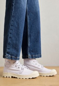 White canvas high-top sneakers with a textured rubber sole, featuring metal eyelets, and a circular logo patch on the side.