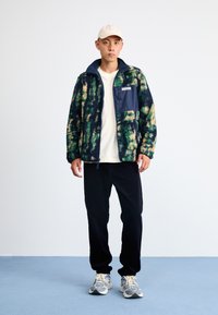 Fleece striukė su žalia, tamsiai mėlyna ir geltona rašte, su tamsiai mėlyna krūtine kišenėje, dėvima ant baltos marškinėlių, derinama su juodomis kelnėmis ir sportbatiais.