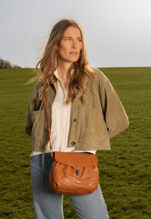Frau mit langen Haaren, die eine beige Jacke, ein weißes Hemd, hellblaue Jeans und eine braune Leder-Umhängetasche trägt, steht auf einem grasbewachsenen Feld.