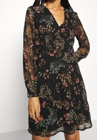 Schwarzes Blumenkleid mit langen transparenten Ärmeln, tiefem V-Ausschnitt und einem fließenden Schnitt mit bunten Blumenmustern und leichter Textur.