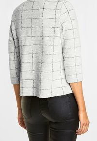 Vit texturerad jacka med svart rutmönster och trekvartslånga ärmar, bärs med svarta byxor och fotograferad bakifrån.