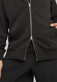 Sort zip-up hættejakke lavet af blødt stof, med en sølvfarvet lynlås, to forlommer og ribbede manchetter. Bæres over matchende sorte bukser.