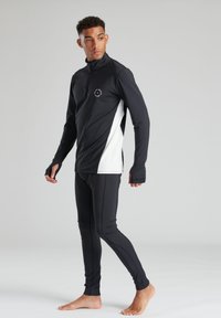 Zwart-witte sporttop met een kwart rits, duimgaten en een logo op de borst, gecombineerd met bijpassende zwarte strakke leggings.