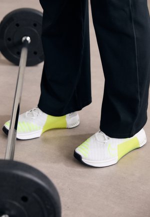 Personne portant des baskets blanches et jaune fluo et un pantalon noir, debout près d’une barre avec des poids sur le sol d’une salle de sport.