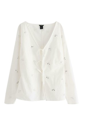 Bílá blůza s dlouhým rukávem a výstřihem do V, zdobená květinovou výšivkou a korálkovými detaily. Lehce pružný materiál s zavazováním v přední části.