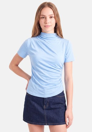 Ung kvinde med langt lyst brunt hår bærer en lyseblå t-shirts med korte ærmer og en mørk denim nederdel, stående mod en hvid baggrund.