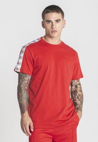 Mladý muž s krátkymi vlasmi, oblečený v červenom tričku s bielymi vzorovanými pruhmi na rukávoch a červených nohaviciach, s rukami čiastočne v vreckách.