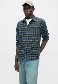 Πουκάμισο φλάνελα σε καρό σχέδιο σε ναυτικό μπλε, πράσινο και ανοιχτό μπλε με κουμπιά μπροστά, τυλιγμένα μανίκια και λευκή μπλούζα με λαιμόκοψη από κάτω. Φορεμένο με ανοιχτόχρωμα τζιν.