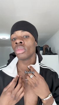 Casaco atlético preto com colarinho branco, colar em camadas com cruz prateada, anéis prateados e um durag preto. O fundo apresenta sapatos e roupas.