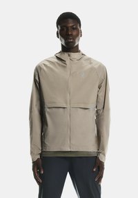 Beige vieglais apģērbs ar kapuci, ar priekšējo rāvējslēdzēju, atstarotājiem un brīvu piegriezumu, izgatavots no gluda, ūdensizturīga materiāla.