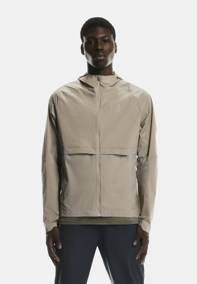 Beige vieglais apģērbs ar kapuci, ar priekšējo rāvējslēdzēju, atstarotājiem un brīvu piegriezumu, izgatavots no gluda, ūdensizturīga materiāla.