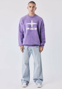 Sudadera de forro polar morada con el logotipo "PXP" en blanco, combinada con jeans holgados de lavado claro y zapatillas blancas. El modelo lleva una cadena de plata y anillos.