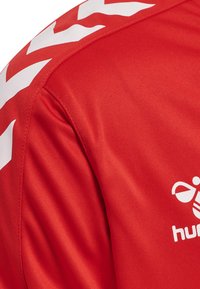 Rode sporttrui van stof met witte chevronpatronen op de schouders en een geprinte logo. Soepele textuur met zichtbare stikseldetails.