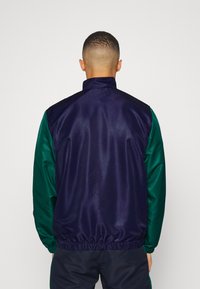 Bomber jacket s barevnými bloky v námořnické modré a zelené barvě, s vysokým límcem, elastickým pasem a hladkou saténovou texturou. Zadní pohled ukazuje kontrastní rukávy.