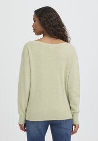 Pull surdimensionné vert clair avec col et poignets côtelés, présentant une texture douce. Porté avec un jean bleu, vu de dos.