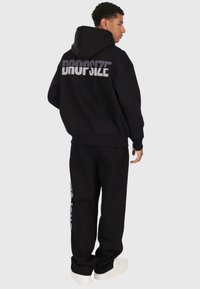 Schwarzer Kapuzenpullover mit großem, silbernem "DROPSIZE"-Druck auf der Rückseite, kombiniert mit schwarzen Jogginghosen. Weicher, lässiger Stoff.