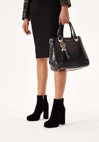 Botas de tobillo de ante negro con un tacón grueso y cremalleras laterales, combinadas con un bolso negro texturizado que presenta detalles dorados y un adorno de borlas.