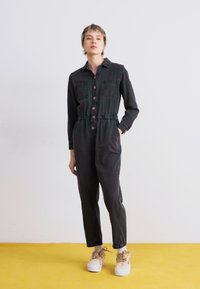 Jumpsuit in denim nero con bottoni frontali, maniche lunghe, vita elastica e tasche laterali. Indossato con sneakers platform beige chunky su uno sfondo giallo.