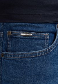 Vaqueros de mezclilla azul con un bolsillo frontal que tiene una etiqueta plateada que dice "PETROLIND". Con costuras distintivas y un diseño de corte clásico.