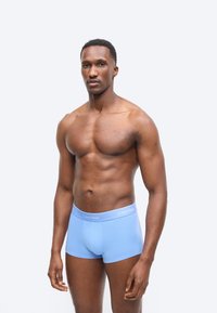 Μυώδης άντρας όρθιος, φορώντας ανοιχτό γαλάζιο μποξεράκι Calvin Klein, μπροστά σε μια απλή ανοιχτογκρίζα φόντο.