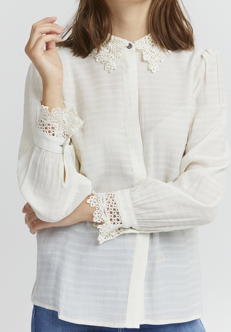 Femme portant un chemisier blanc transparent avec col et poignets en dentelle, debout les bras croisés et une main levée près du visage.