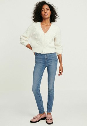 Vit stickad kofta med V-ringning och texturerad flätmönster, matchad med ljusblå slim fit jeans och rosa volangskor.