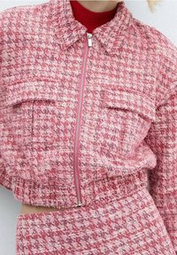 Femme portant une veste zippée en tweed rose et blanche avec de grandes poches avant, sur un haut rouge et une jupe en tweed assortie.