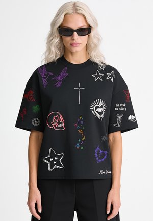 Femme aux longs cheveux blonds et lunettes de soleil noires portant un T-shirt noir oversize avec des imprimés graphiques colorés et un texte "no risk no story".