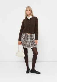 Καφέ κοντό καρντάν με κούμπωμα, καρό μίνι φούστα σε μπεζ και καφέ αποχρώσεις, μαύρες καλσόν και μαύρα φλατ παπούτσια. Μικρή λευκή τσάντα.