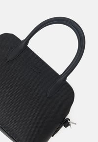Schwarze strukturierte Handtasche mit glatten Ledergriffen, Reißverschluss und einem kleinen Logodetail auf der Vorderseite. Kompaktes rechteckiges Design.
