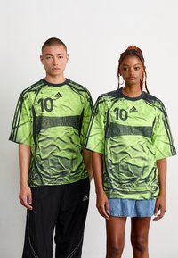 To modeller iført overdimensjonerte grønne og svarte Adidas sportstrøyer med nummer 10, stående side om side mot en ensfarget bakgrunn.