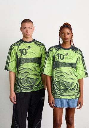 To modeller iført overdimensionerede grønne og sorte Adidas sports trøjer med nummer 10, står side om side mod en ensfarvet baggrund.