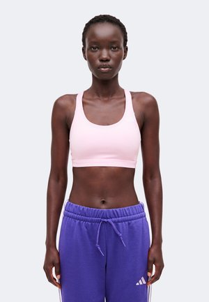 Mladá osoba nosiaca svetloružovú športovú podprsenku a fialové tepláky Adidas stojí pred jednoduchým svetlým pozadím.