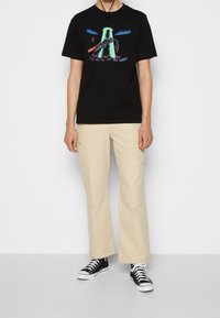 Svart bomull T-shirt med färggrafik som visar en hand som håller i en kedja, kombinerad med beige cargo-byxor och svarta sneakers.