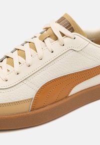Beige and brown leather sneaker with textured tan tongue, cream laces, stitched logo stripe, and gum rubber sole against white background.