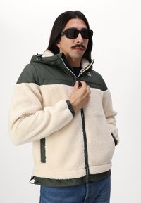 Giacca in pile con un corpo morbido di colore crema e una parte superiore verde scuro, cappuccio regolabile, polsini elasticizzati, chiusura con zip e tasche laterali.
