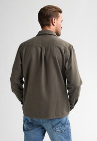 Olive green long-sleeve shirt with a collar, buttoned cuffs, and a back yoke detail, paired with blue denim pants. Soft fabric texture.