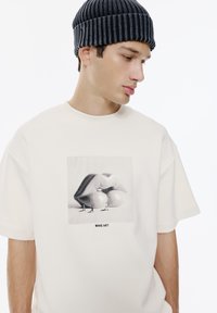 Maglietta oversize color crema con una grafica di labbra lucide sopra il testo "MAKE ART." Il modello indossa un berretto nero a coste.