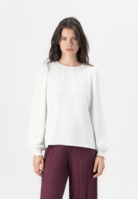 White long-sleeve blouse with textured fabric, round neckline, and gathered accents at the cuffs, paired with pleated burgundy trousers.