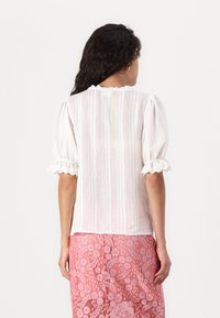 Blouse rayée blanche avec des manches bouffantes et des poignets volants. Présente un décolleté festonné. Portée sur une jupe en dentelle rose avec des motifs floraux.