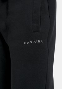 Svarta joggerbyxor i mjukt material, med slim fit, sidofickor och en diskret "CASPARA"-logga i grått på lårområdet.