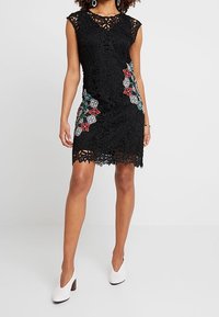 Robe en dentelle noire avec manches courtes, broderies florales en rouge et bleu, ourlet festonné et tissu texturé. Portez avec des talons blancs.