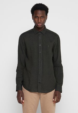 Ung mand iført en langærmet mørkegrøn skjorte med knapper og beige bukser, stående mod en ensfarvet lys baggrund.