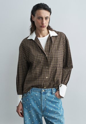 Camisa de cuadros marrón oversize con cuello blanco, combinada con jeans azules que tienen detalles en perlas blancas y acentos dorados.