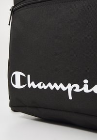Schwarzer Stoff-Rucksack mit strukturierter Oberfläche, auf dem ein großes, weißes "Champion"-Logo deutlich sichtbar ist. Der Reißverschluss ist oben sichtbar.