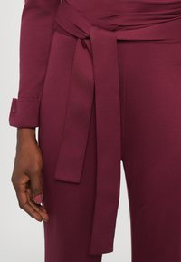 Burgundska obleka s preklopnim dizajnom, s detajlom vezanim okoli pasa. Tkanina ima gladko teksturo, srajca pa ima manšete na gumb.