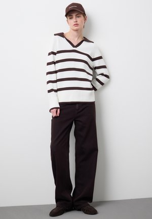 Personne portant un pull blanc à rayures horizontales foncées, un pantalon large foncé, des chaussures foncées assorties et une casquette de baseball foncée, se tenant contre un mur blanc.