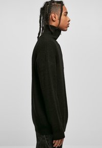 Pull en laine noir à col roulé avec une texture côtelée, doté de manches longues et d'une coupe décontractée, présenté en vue de profil sur un fond neutre.