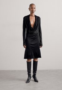 Rochie neagră, cu mânecă lungă, cu un front drapat și tiv până la genunchi. Textură mată. Asortată cu cizme stiletto până la genunchi.