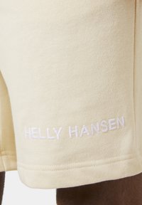 Crème-kleurige shorts gemaakt van zachte stof, met geborduurd "HELly HANSEN"-merk in het wit op de zoom, met een gladde textuur.
